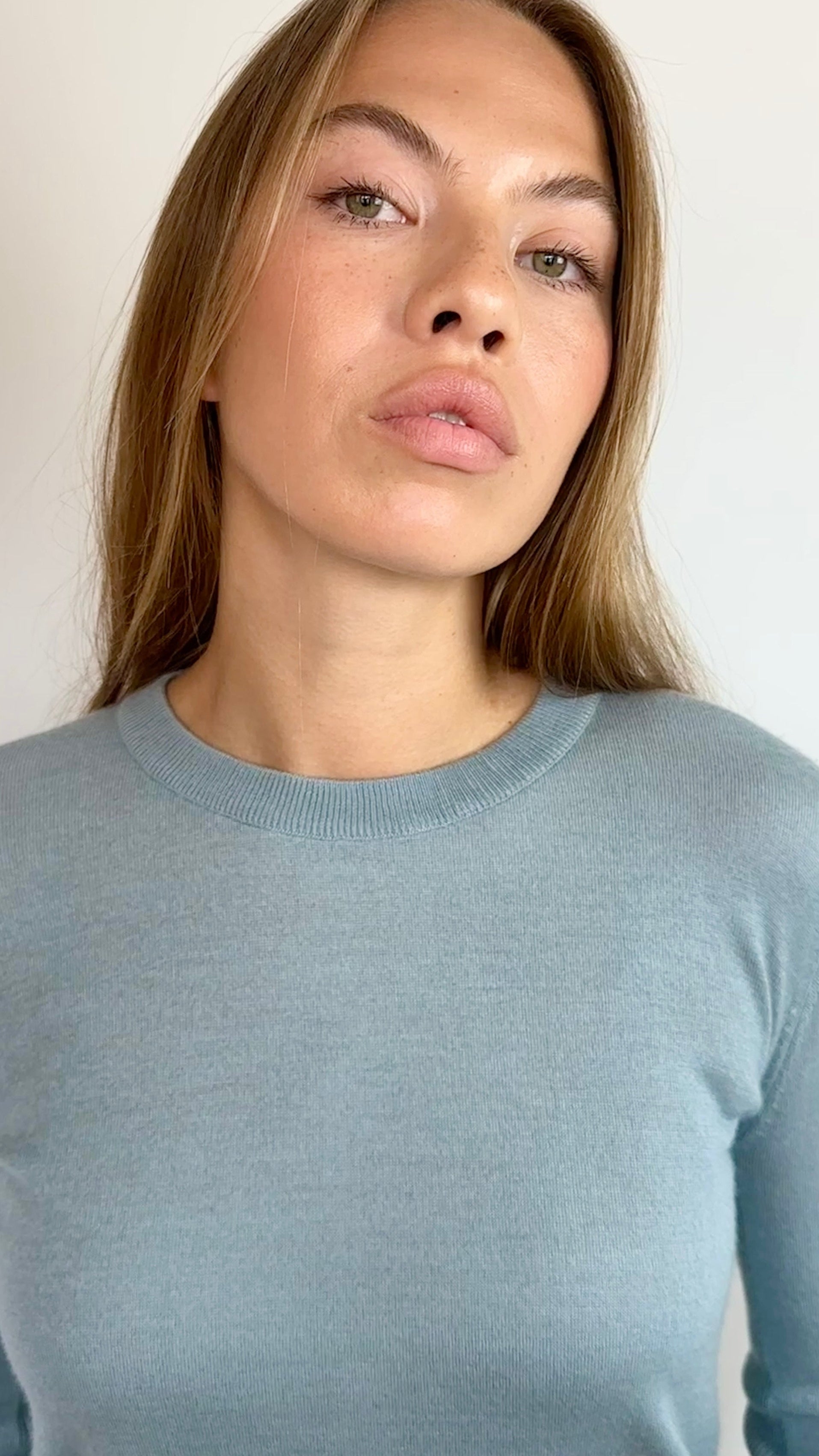 A woman with blonde hair wearing a light blue long-sleeve crewneck knit sweater, photographed against a plain off-white background.