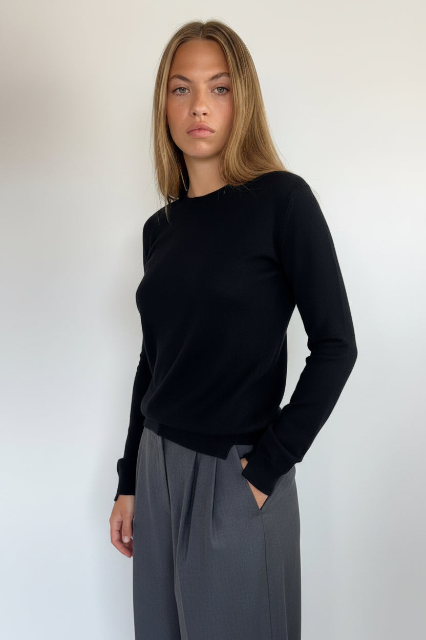 A woman modeling a black long-sleeve knit sweater with dark gray draped trousers, standing against a plain off-white wall.