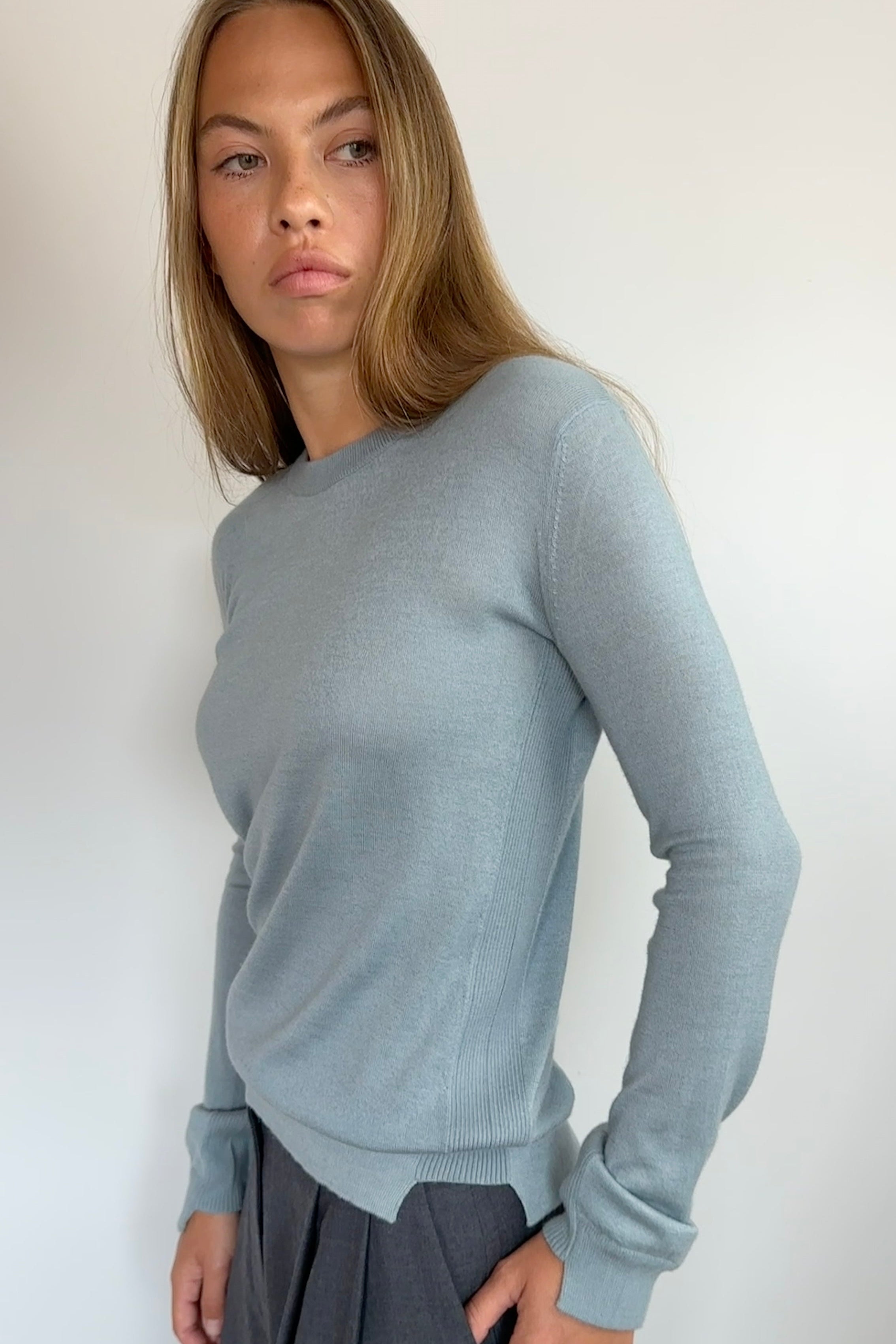 A woman modeling a light blue-gray long-sleeve knit sweater with dark gray draped trousers, standing against a plain off-white wall.