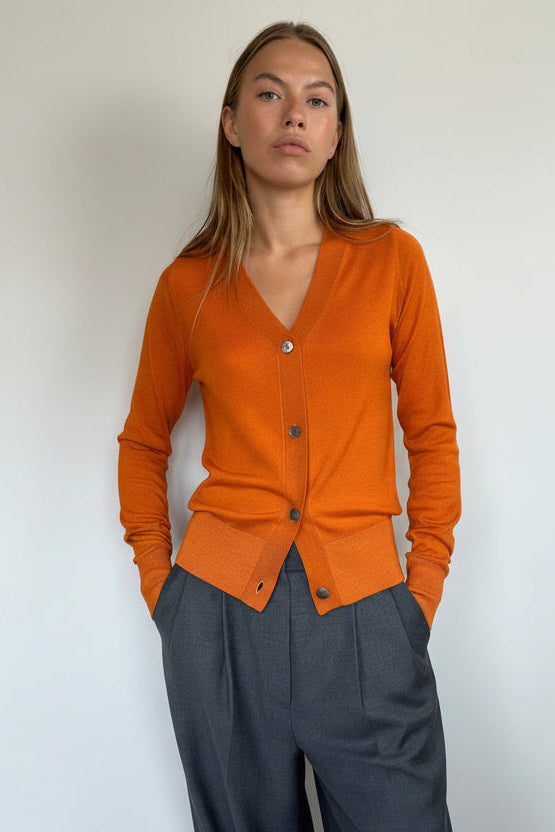 A woman modeling a bright orange long-sleeve button-front cardigan with a V-neck, paired with dark gray wide-leg pants, standing against a plain light gray wall.