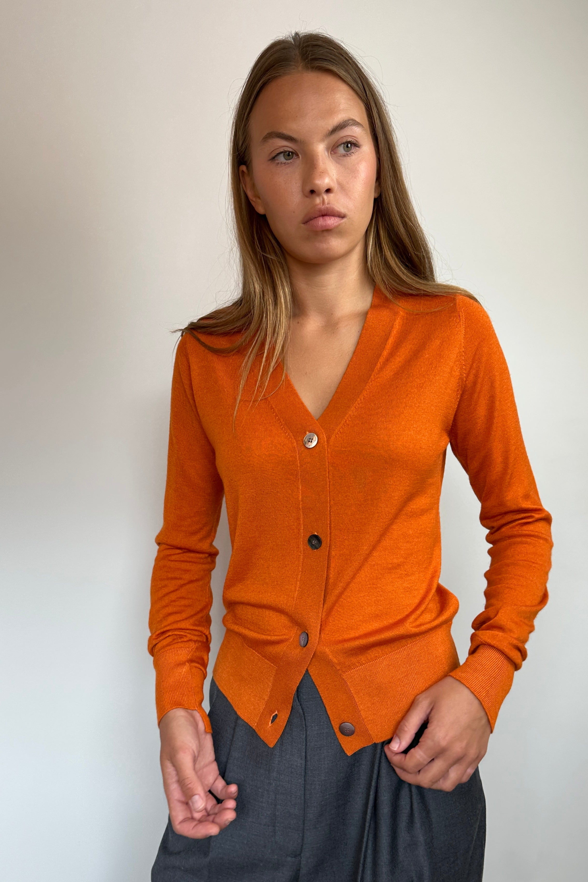 A woman modeling a rust-orange long-sleeve button-up cardigan sweater paired with dark gray wide-leg pants, standing against a plain off-white wall.