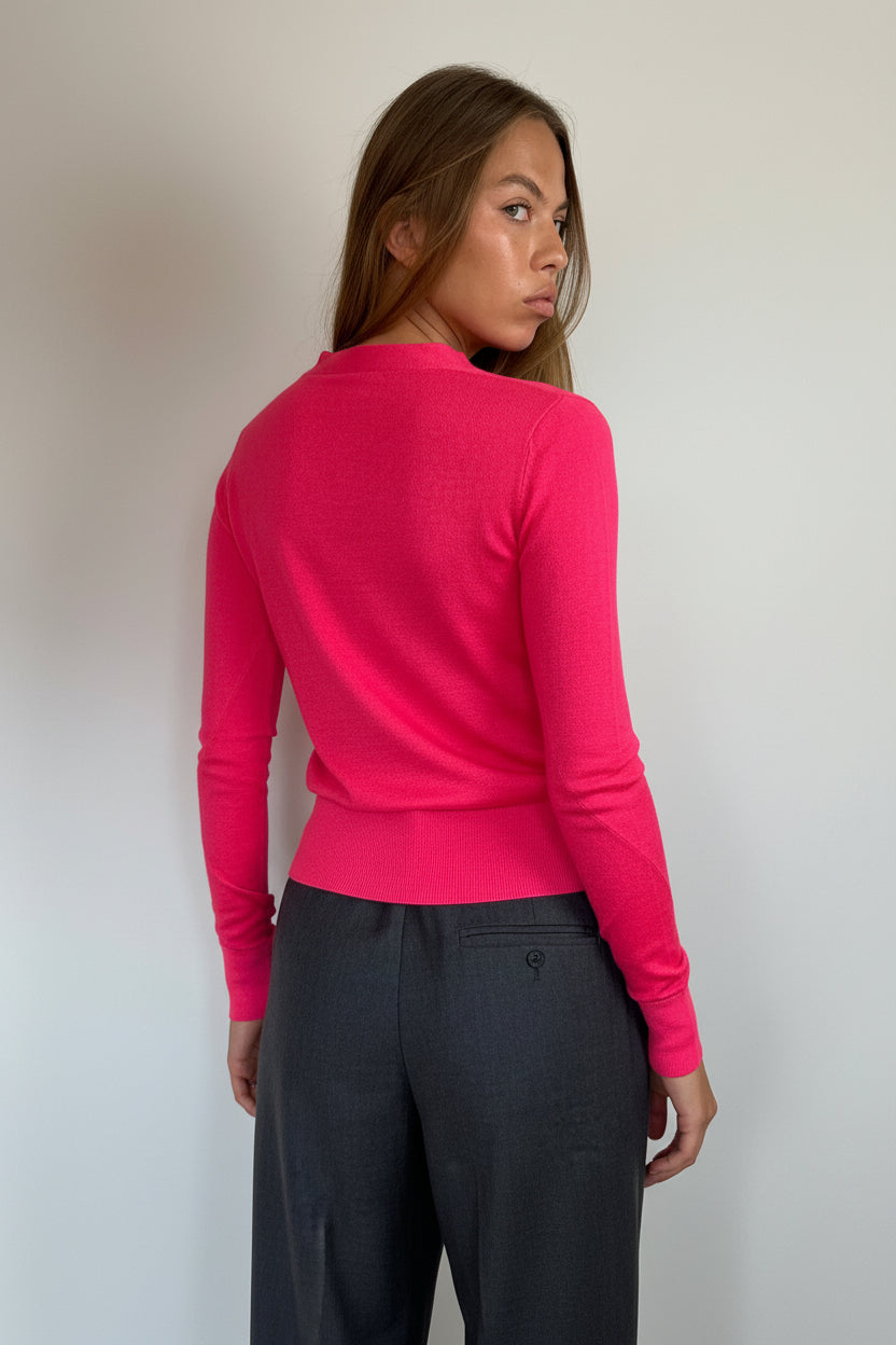 A woman wearing a bright pink long-sleeve knit top and dark gray trousers, photographed from the back against a plain light gray wall.