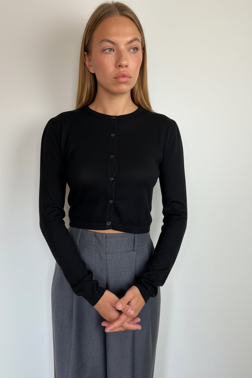 A woman modeling a black long-sleeve cropped button-up top paired with gray trousers, standing against a plain light gray wall.