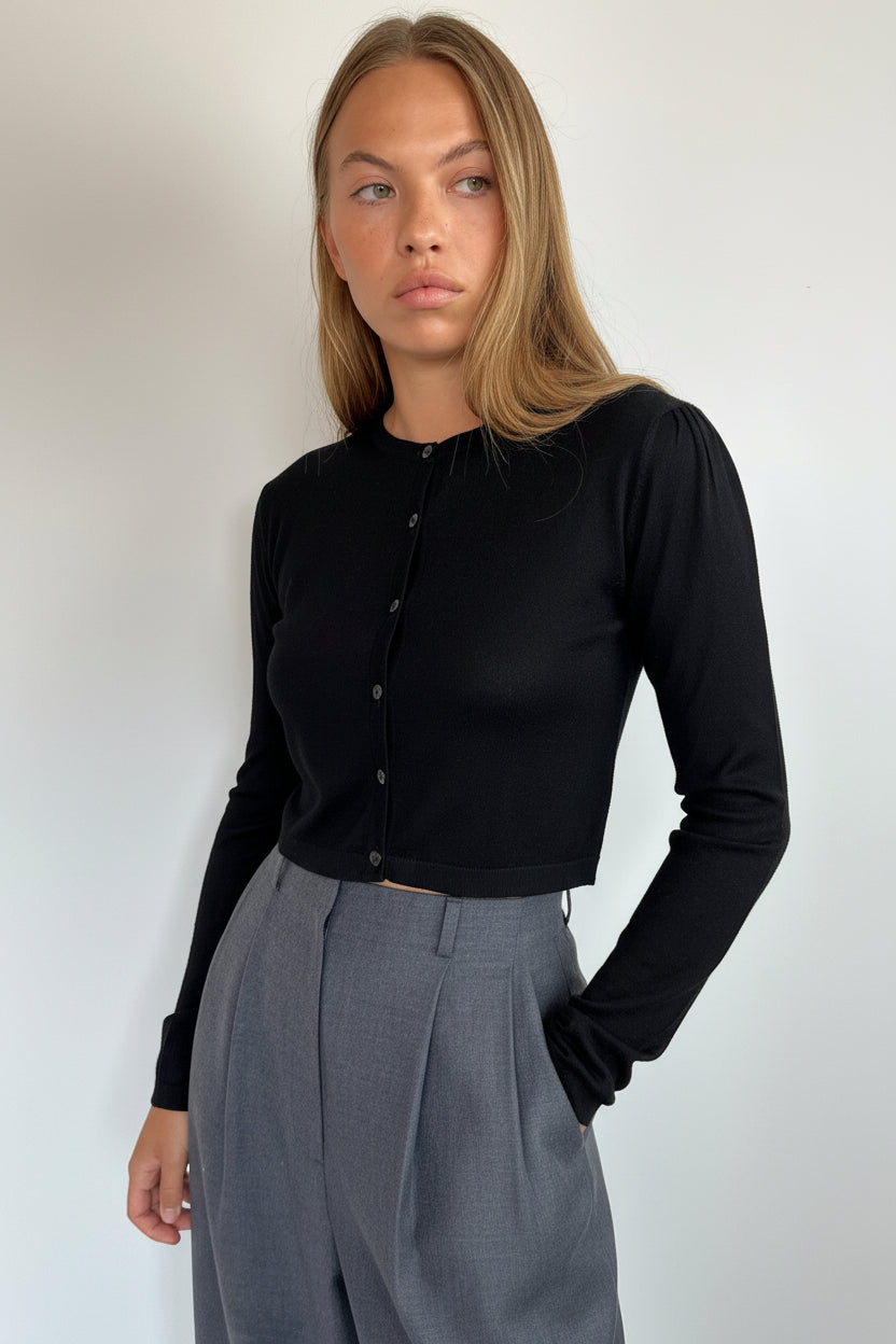 A woman wearing a black long-sleeve button-front top and gray trousers, standing against a plain light gray wall.