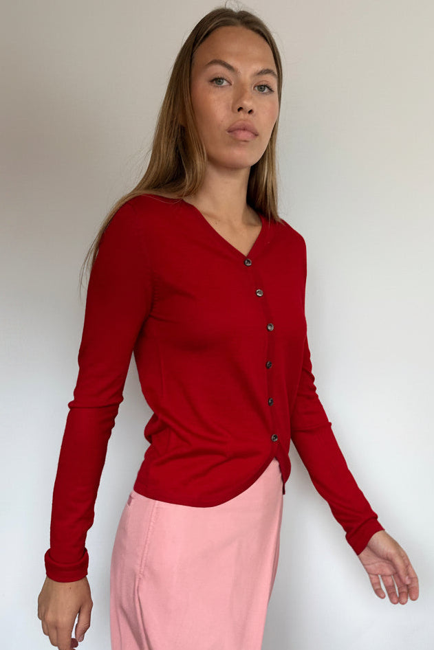 A woman modeling a red long-sleeve button-front cardigan over a light pink skirt, standing against a plain white wall.