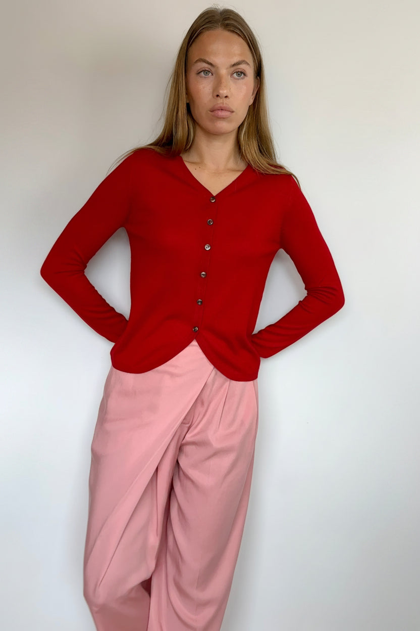 A woman modeling a red long-sleeve button-front cardigan top paired with light pink draped pants, standing against a plain light gray wall.