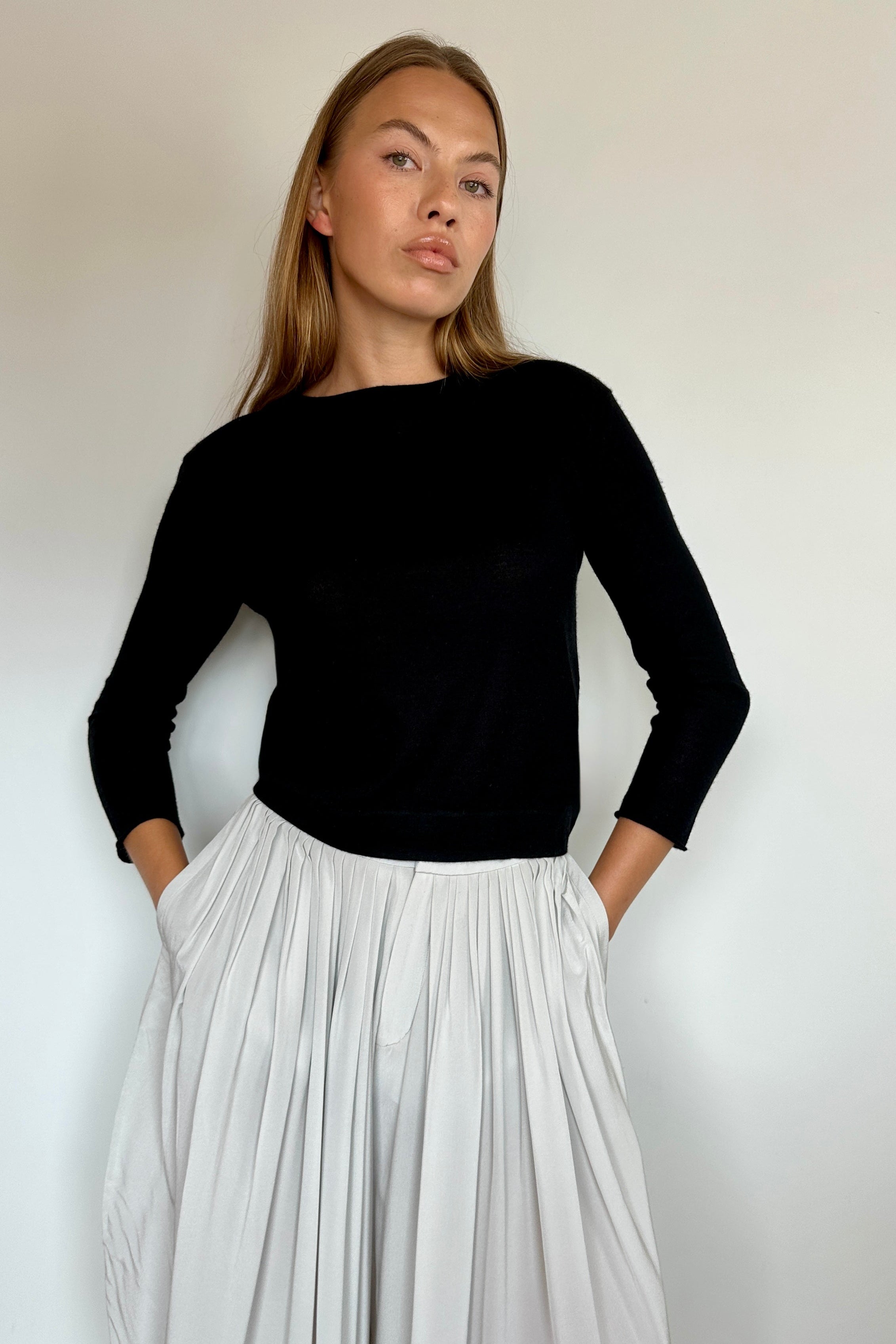A woman modeling a black long-sleeve boat-neck top paired with a high-waisted, pleated white skirt, standing against a plain off-white wall.