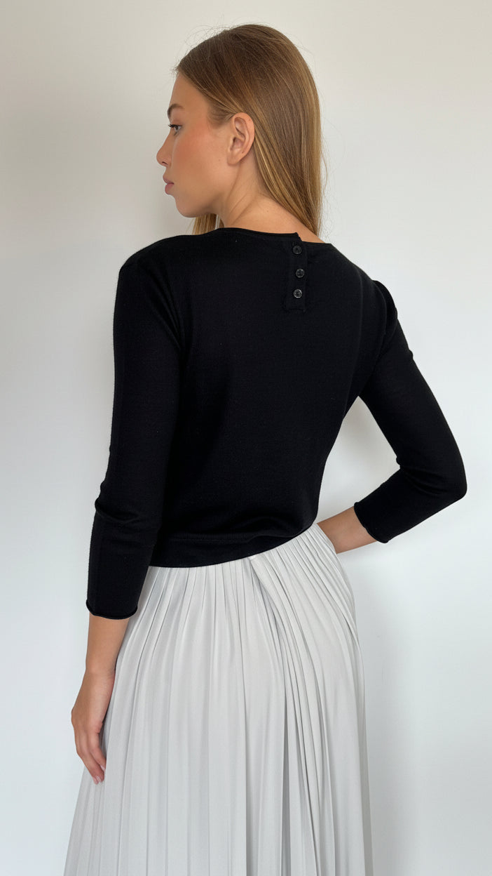 A woman shown from the back wearing a black long-sleeve knit top with three small buttons at the neckline and a light gray pleated skirt, against a plain light gray wall.
