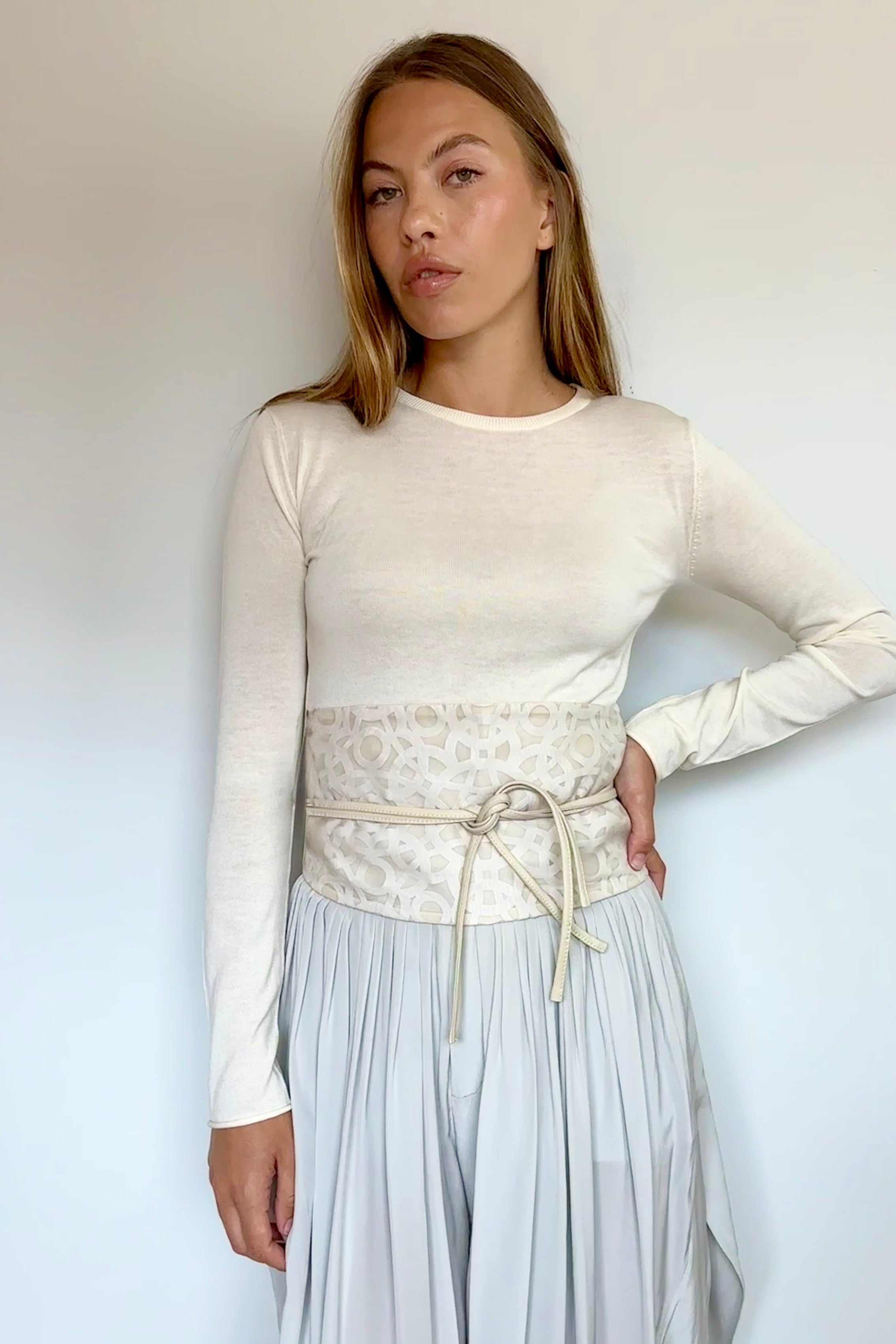 A woman modeling a cream long-sleeve knit top with a wide, patterned cream waist belt tied at the hips, paired with light gray wide-leg pleated pants, against a plain off-white wall.