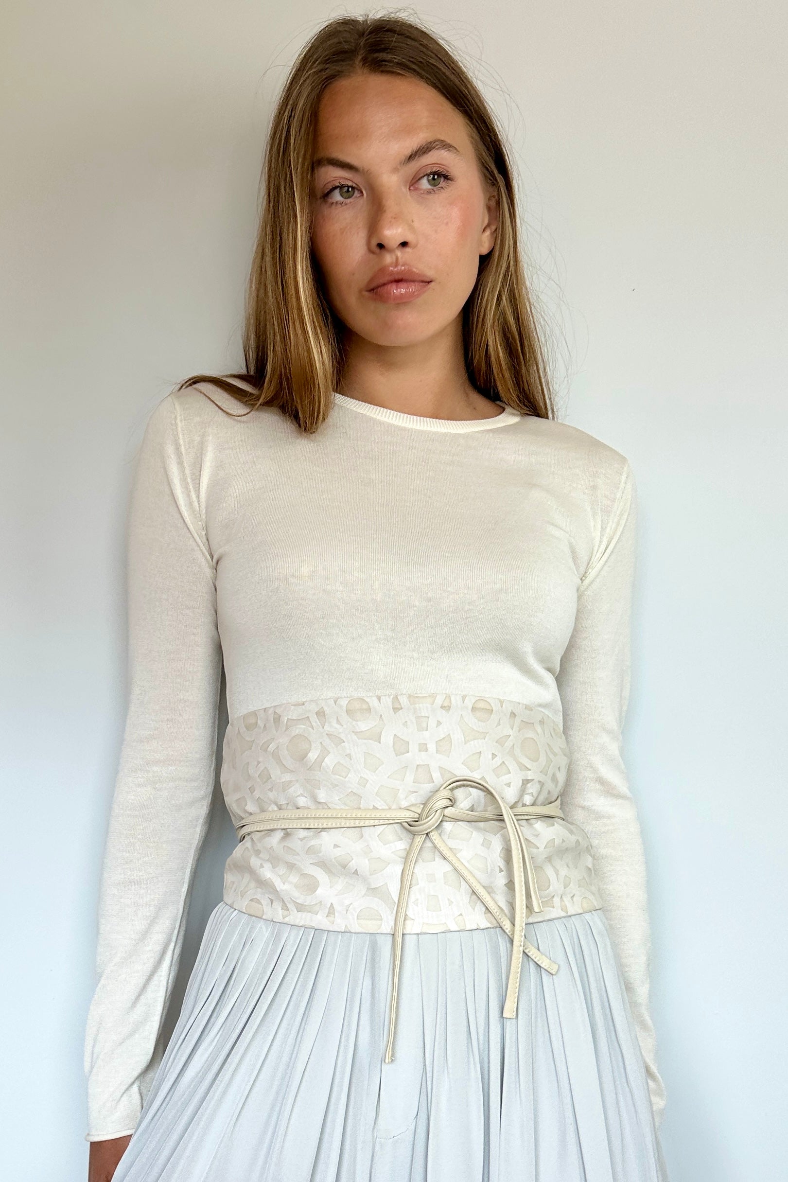 A woman wearing a cream-colored long-sleeve knit top with a textured lace waist panel and a matching drawstring belt, paired with a light blue pleated skirt, against a plain white wall.