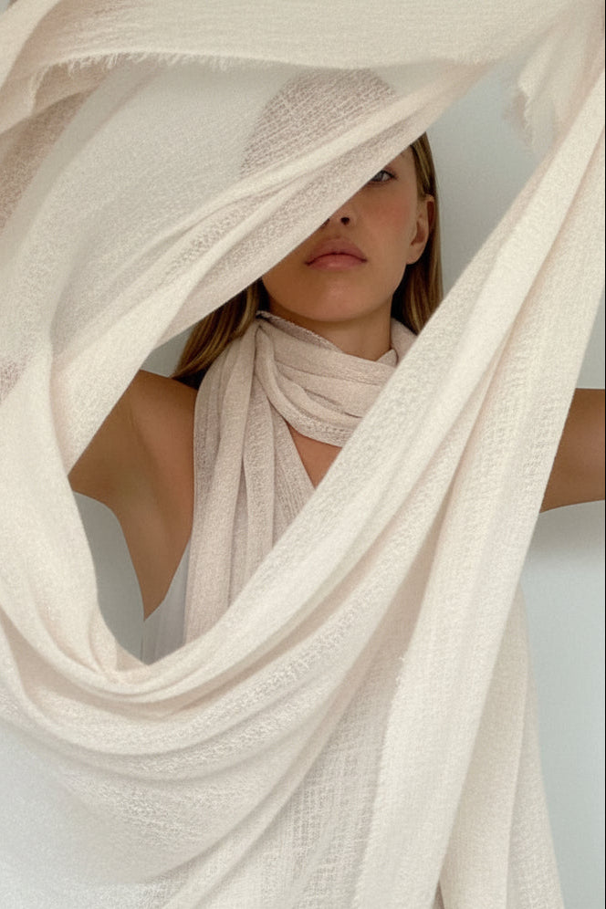 A woman modeling a lightweight, semi-sheer off-white scarf or shawl draped around her shoulders against a plain white wall.