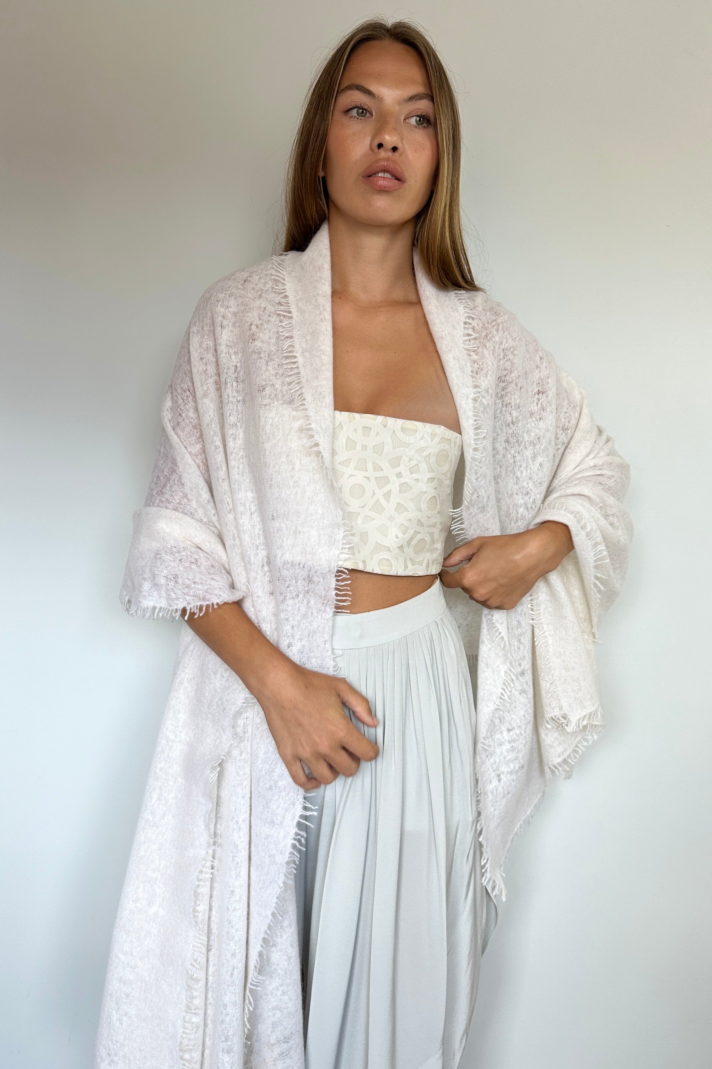 A woman modeling a white lace open-front cardigan over a white strapless crop top and light gray pleated skirt, standing against a plain off-white wall.