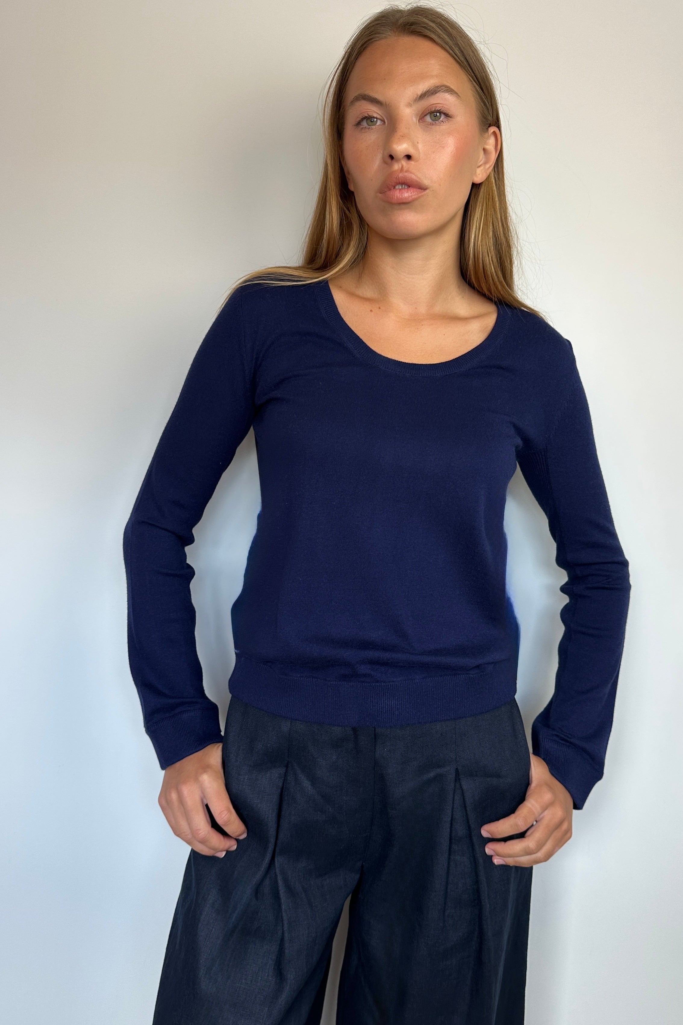 A woman modeling a navy blue long-sleeve knit top with a wide scoop neckline, paired with dark navy wide-leg trousers, standing against a plain off-white wall.