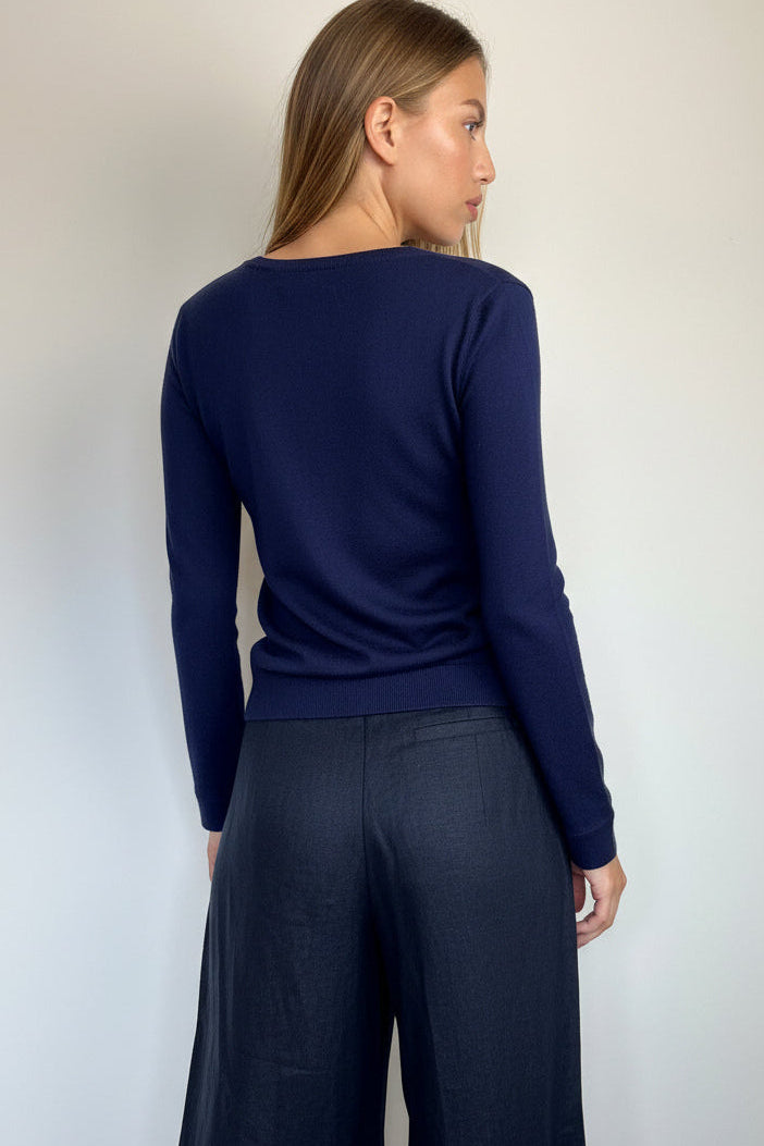 A woman shown from the back wearing a navy blue long-sleeve knit top and dark wide-leg trousers, standing against a plain light gray wall.