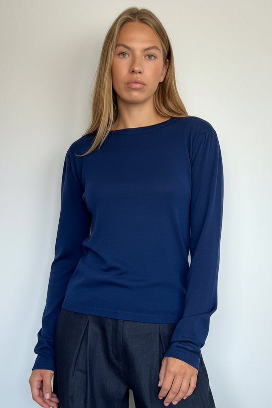 A woman modeling a navy blue long-sleeve boat-neck knit top paired with dark wide-leg trousers, standing against a plain light gray wall.