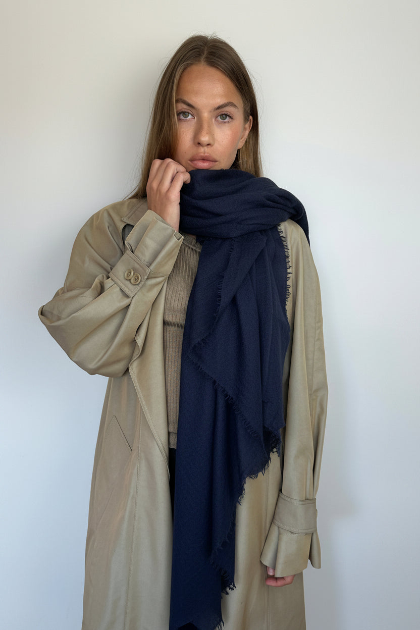 A woman modeling a navy blue scarf wrapped around her neck, paired with a beige trench coat over a ribbed knit sweater, standing against a plain white wall.