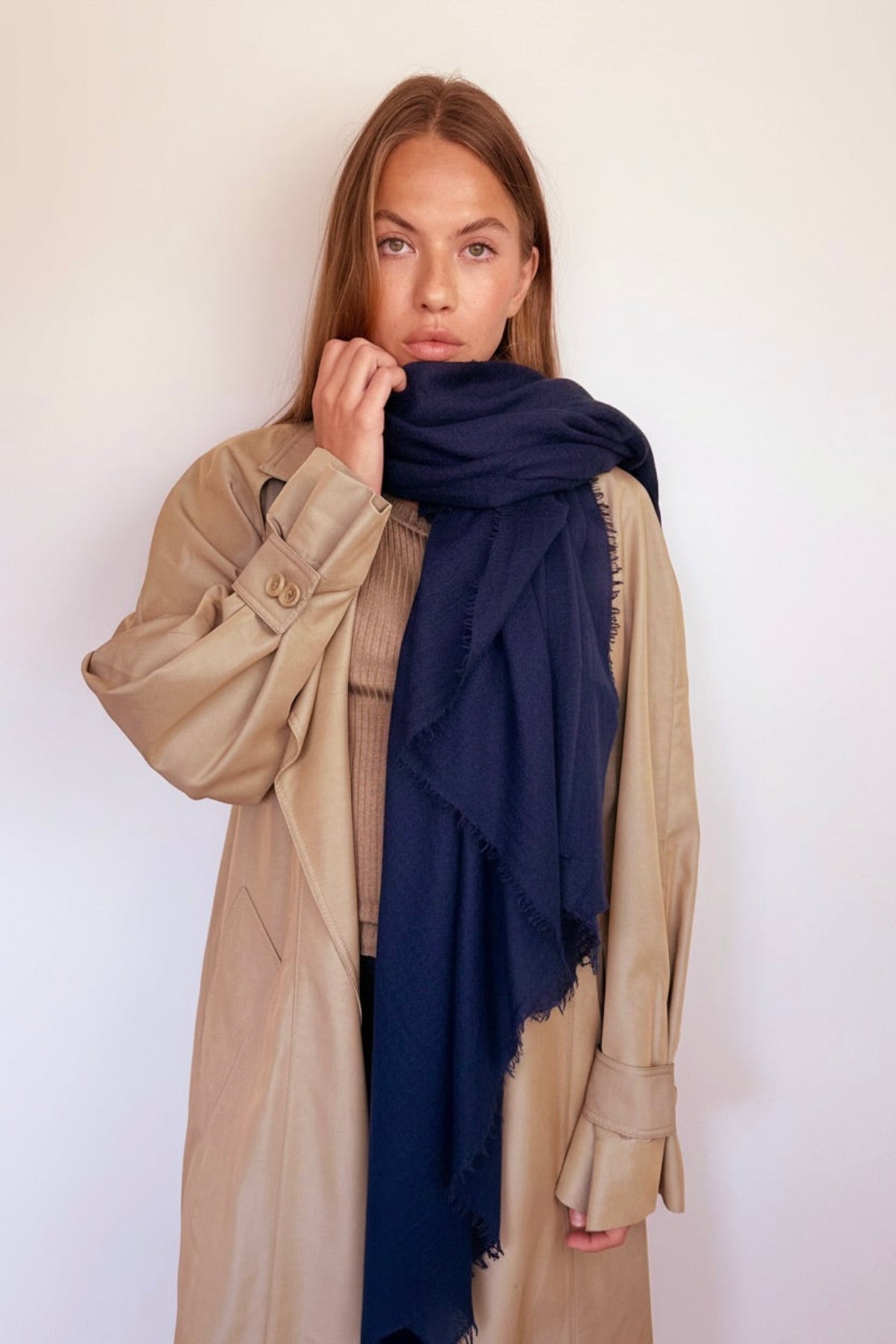 A woman wearing a beige trench coat and a dark navy blue cashmere scarf, posing against a plain off-white background.