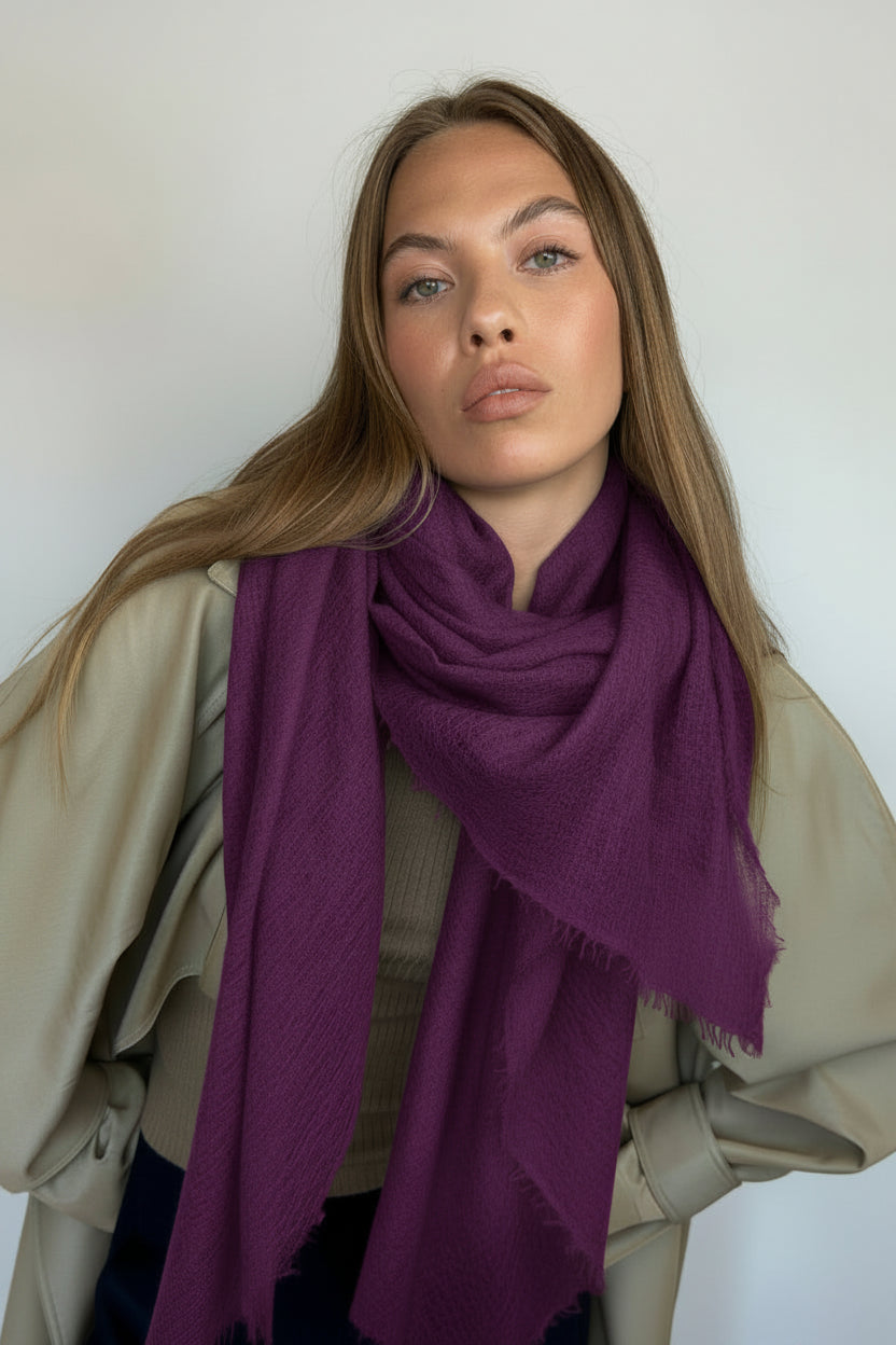 A woman with long light brown hair models a large, deep purple scarf with fringed edges, draped over her shoulders. She is wearing a beige jacket against a plain white wall.