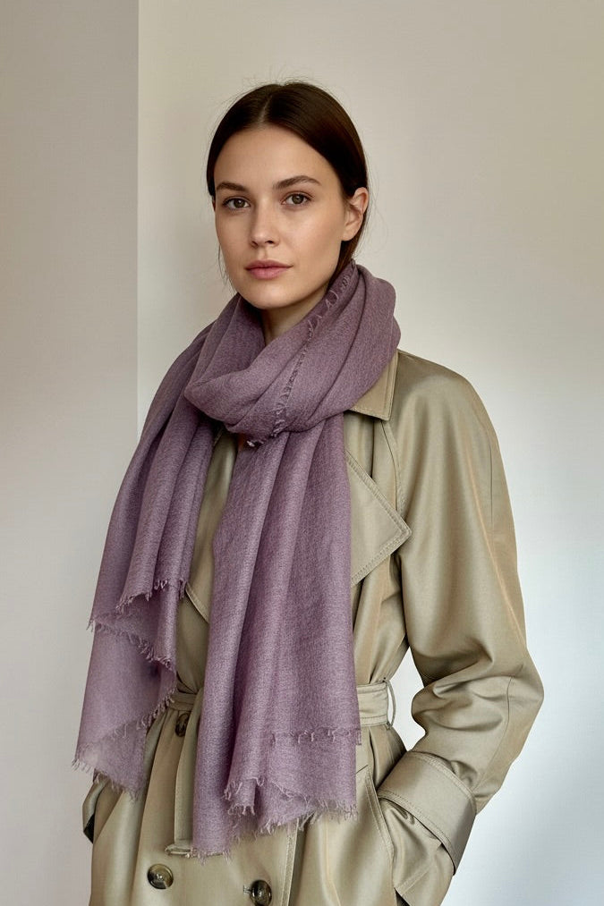 A woman wearing a dusty purple scarf and a beige trench coat, standing against a neutral light gray background.