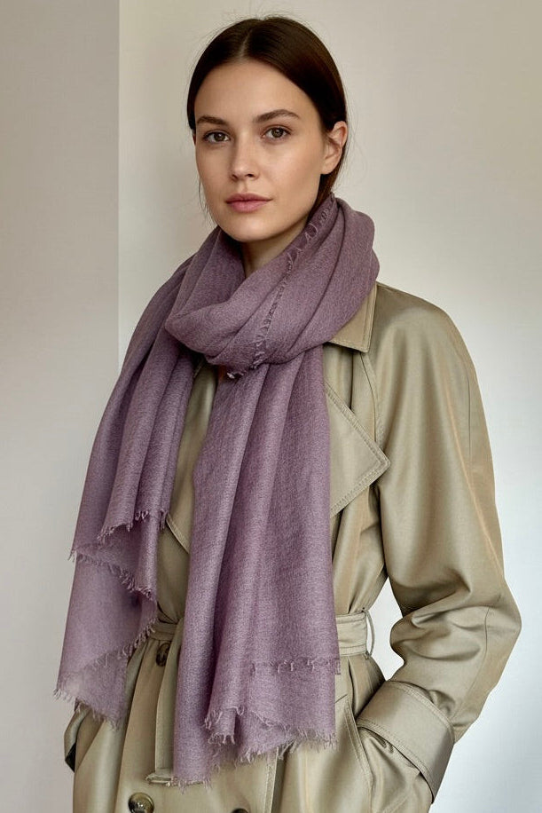 A woman wearing a dusty purple scarf and a beige trench coat, standing against a neutral light gray background.
