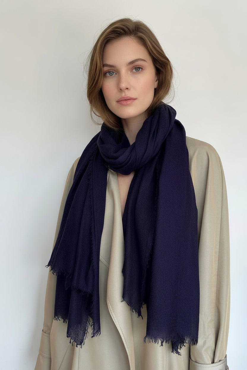 A dark navy blue scarf with fringed edges draped over a beige trench coat on a model against a plain light background.