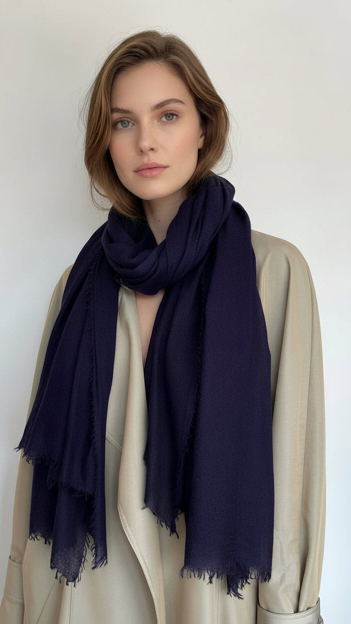 A dark navy blue scarf with fringed edges draped over a beige trench coat on a model against a plain light background.