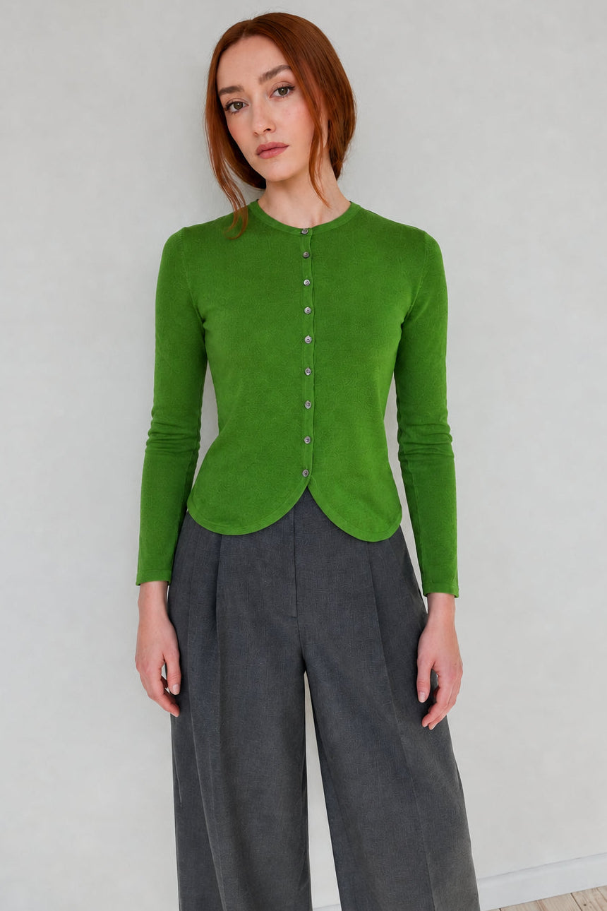 A woman with red hair wearing a bright green long-sleeve button-front top and gray wide-leg pants, standing against a plain light gray wall.