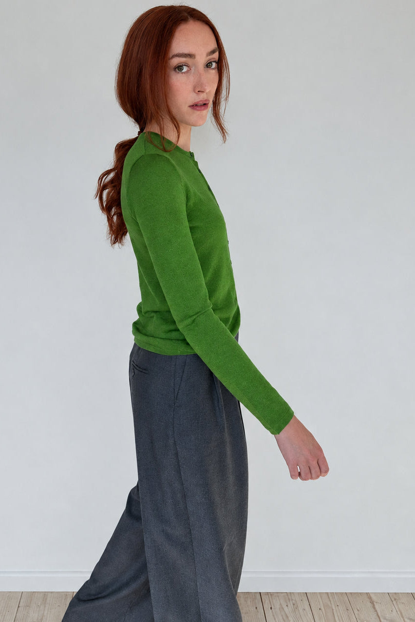 A woman with red hair is shown from the side in a studio setting, wearing a bright green long-sleeve knit top and dark gray wide-leg trousers.