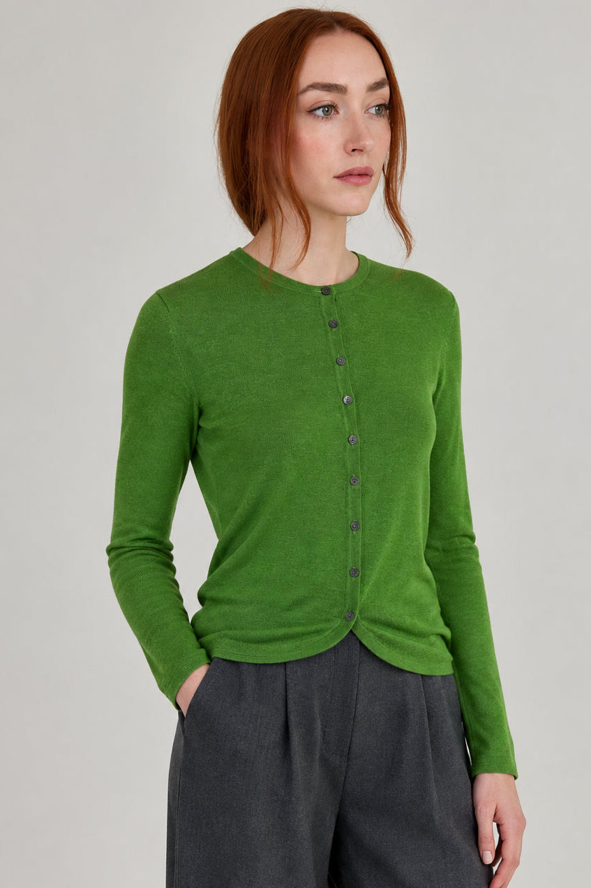 A woman with red hair wearing a bright green long-sleeve button-up cardigan and dark gray pants, standing in a minimalist room with a light gray wall and wooden floor.