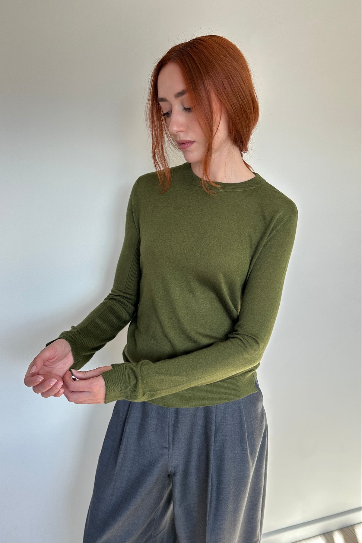 A woman with red hair wearing a long-sleeve olive green knit top and loose gray pants, standing against a plain light gray wall.