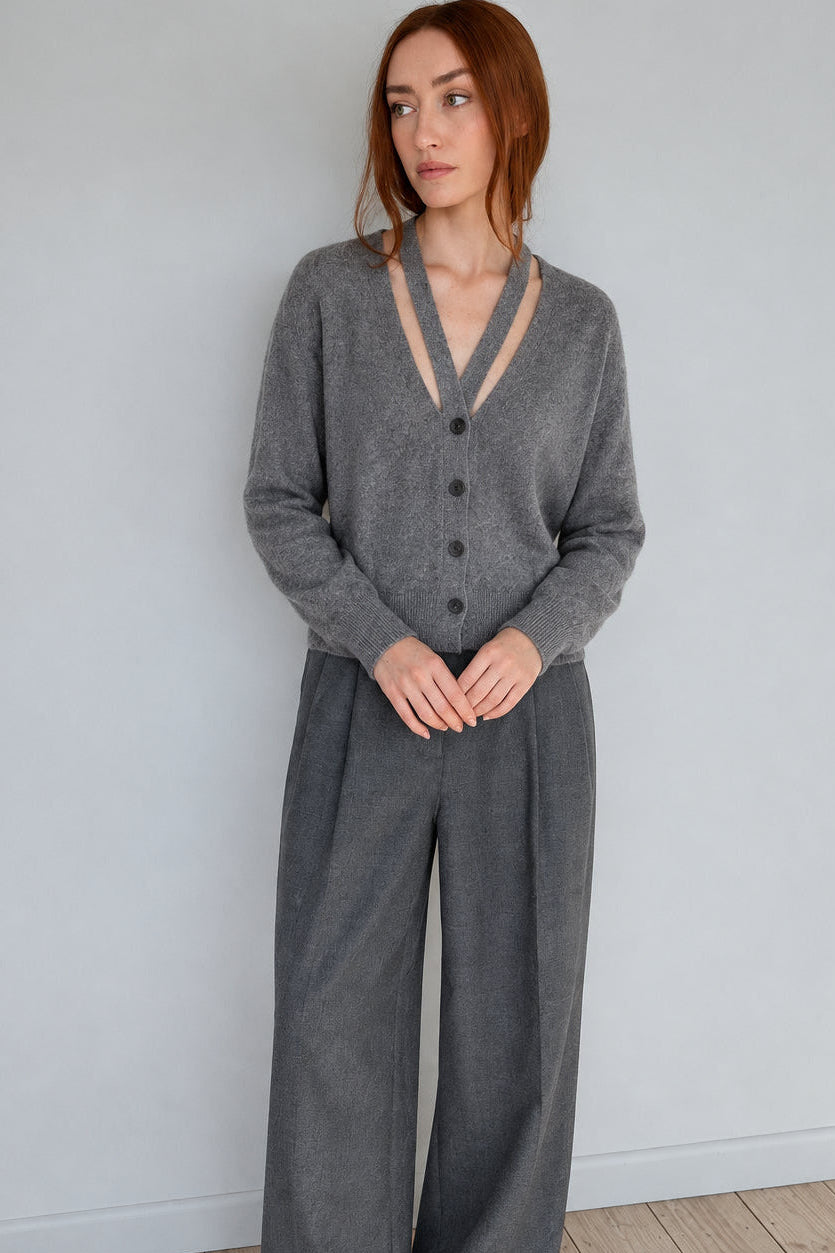 A woman with red hair stands against a plain light gray wall wearing a Marl grey gray button-up cardigan with a beige V-neck trim and wide-leg gray pants.