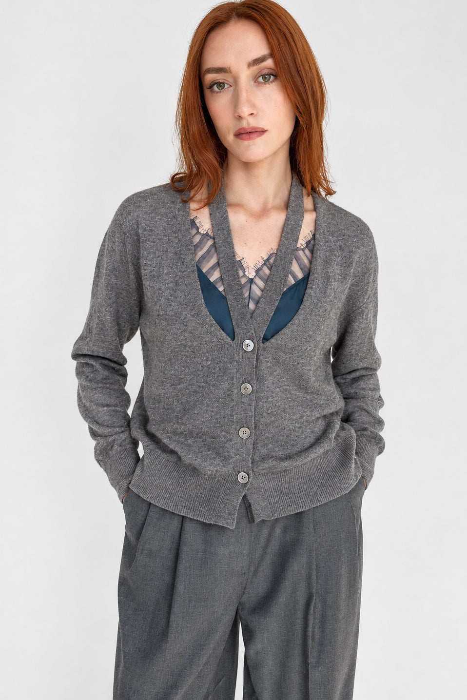 A woman with red hair wearing a mealngee grey gray cashmere button-up cardigan over a blue silk casmisolepaired with gray wide-leg pants, standing against a plain white wall.