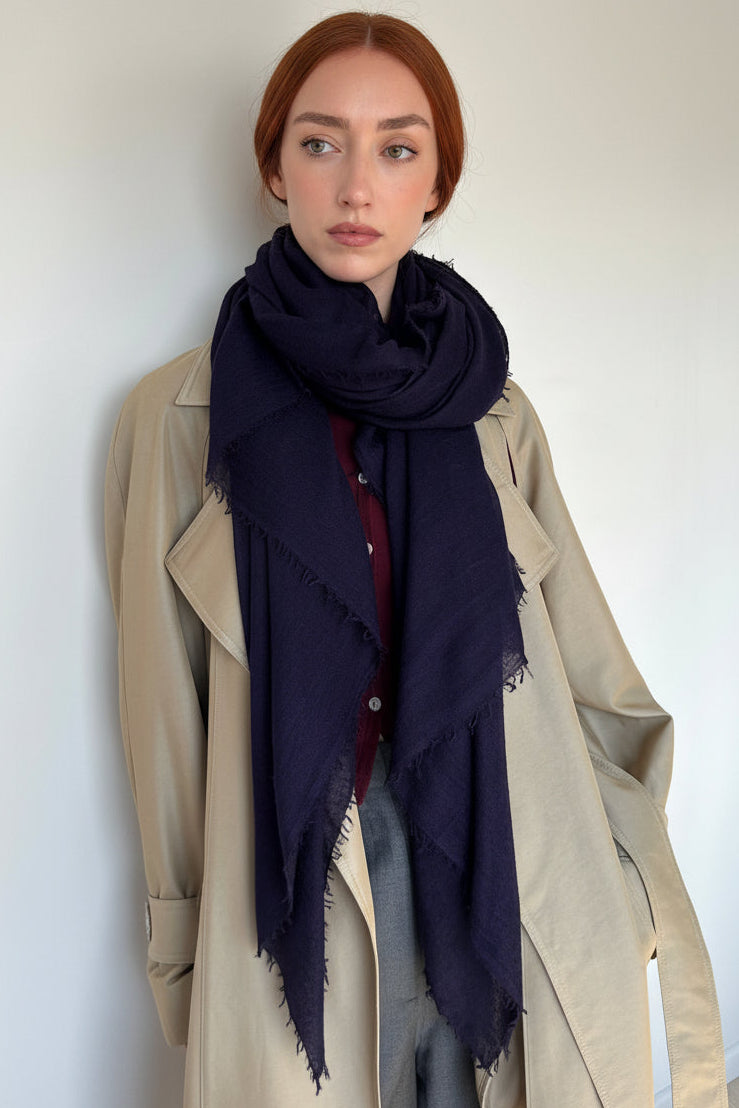 A woman with red hair wearing a beige trench coat, gray pants, and a dark navy scarf with frayed edges, standing against a plain off-white wall.