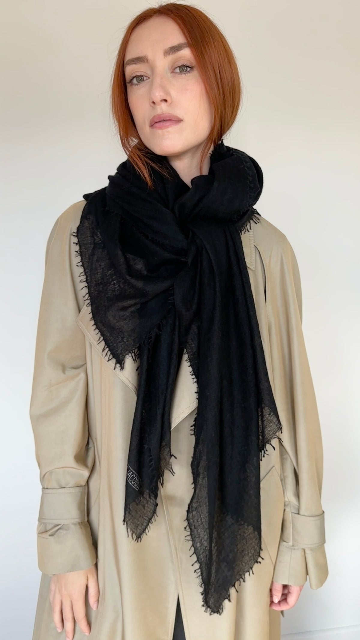 A woman with red hair wearing a beige trench coat and a large black scarf with fringed edges, photographed against a plain light background.