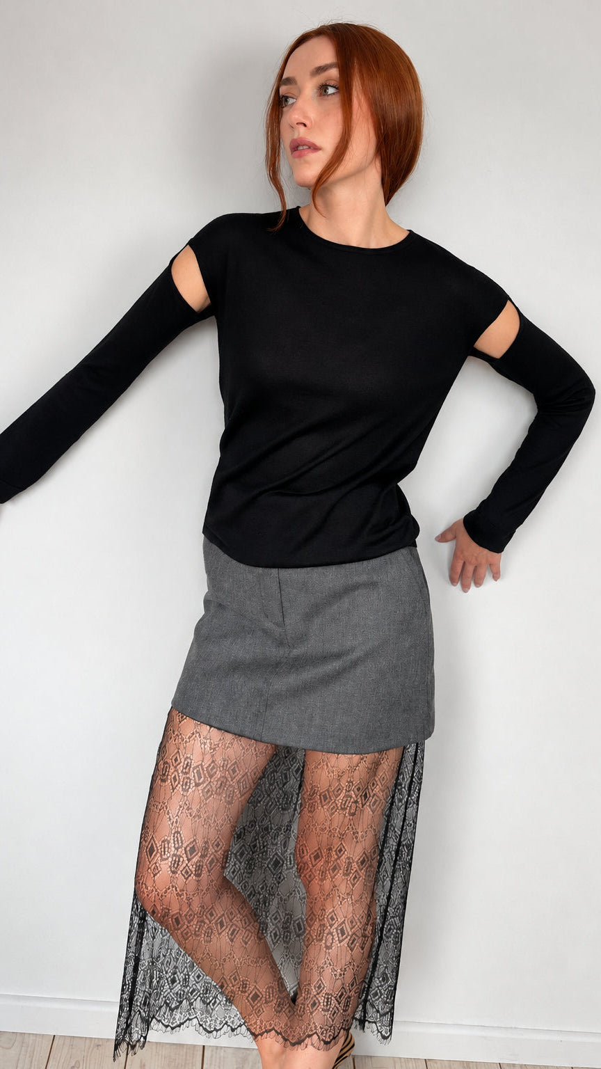 A woman with red hair models a black long-sleeve top with cutout details on the upper arms, paired with a gray mini skirt and sheer black lace tights, standing against a plain white wall.