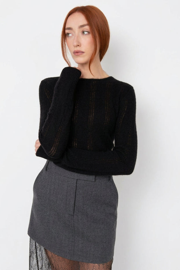 A woman with red hair stands against a plain light gray wall wearing a black ribbed knit long-sleeve top and a gray skirt with a sheer lace overlay.