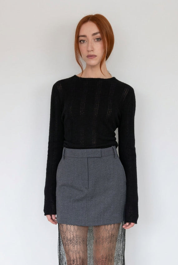 A woman with red hair stands against a plain white wall wearing a black long-sleeve knit top, a gray mini skirt, and sheer black lace tights.