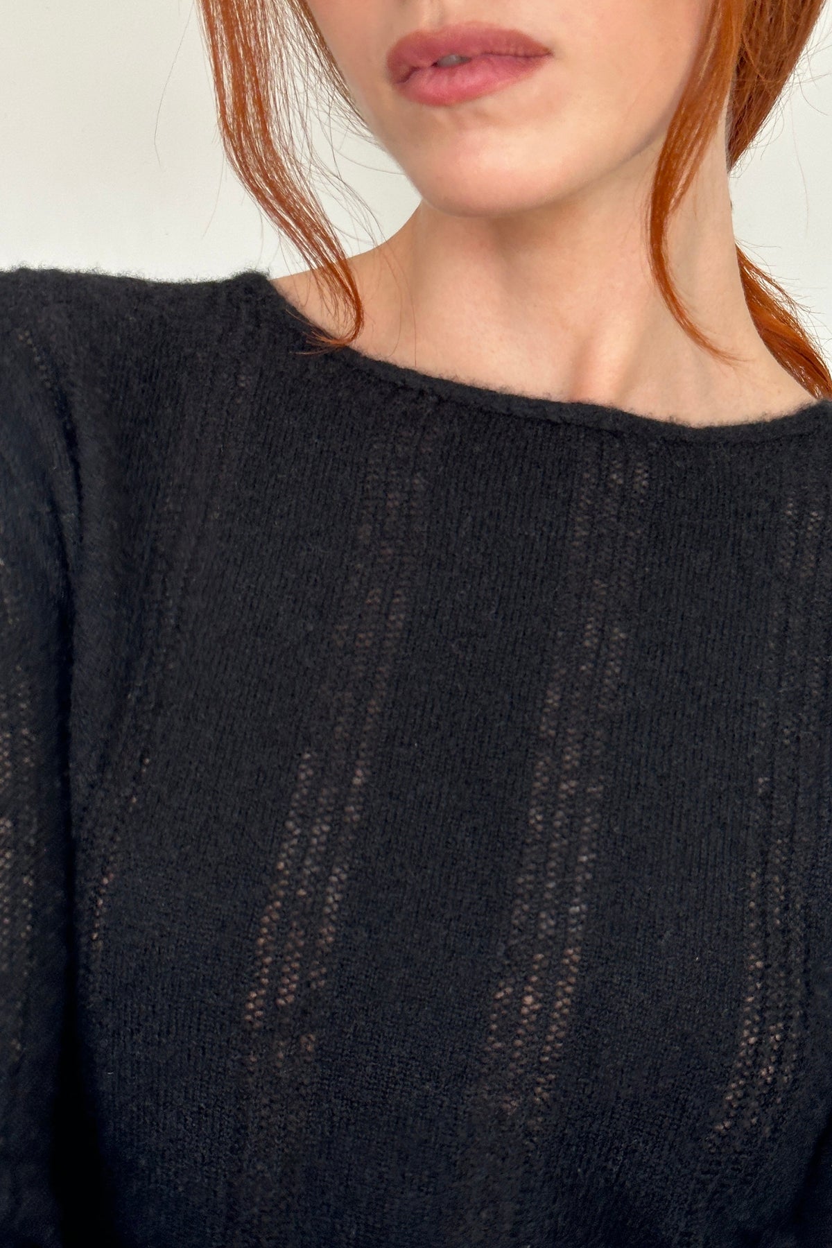 Close-up of a woman with red hair wearing a black open-knit crewneck sweater against a plain light background.