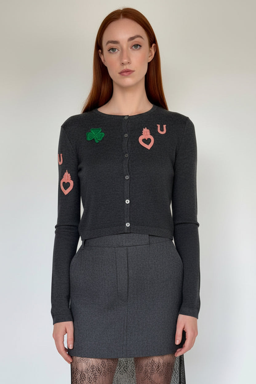 A woman with red hair wearing a dark gray long-sleeve cardigan with green and pink playing card suit embroidery (clubs, hearts, diamonds, kings) on the chest and sleeves, paired with a matching dark gray skirt.