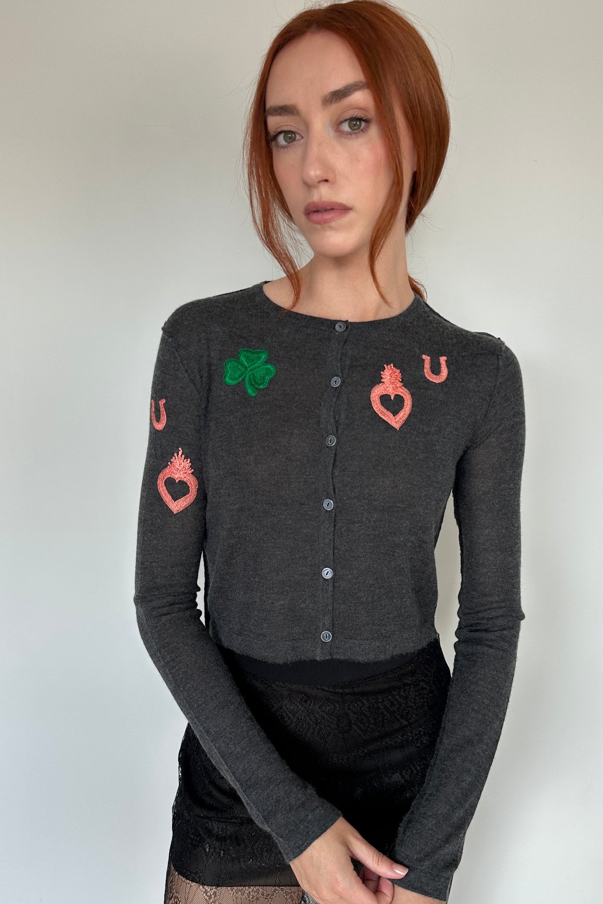A woman with red hair wearing a dark gray long-sleeve button-up cardigan with embroidered playing card suit motifs (green shamrock, red hearts, red horseshoes) on the chest and sleeve, paired with a black skirt.
