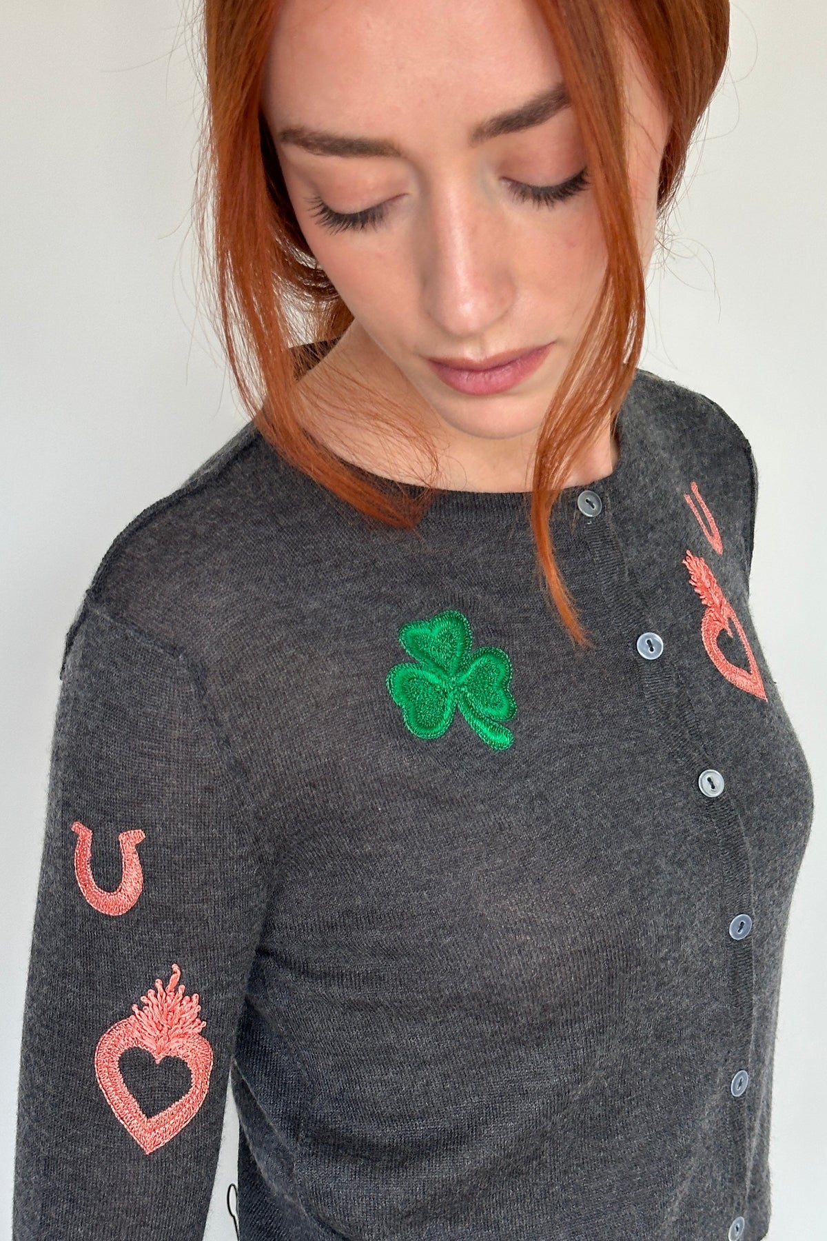 A close-up of a woman with red hair wearing a dark gray long-sleeve button-up cardigan sweater decorated with embroidered St. Patrick's Day motifs, including a green shamrock and pink horseshoe and heart symbols.
