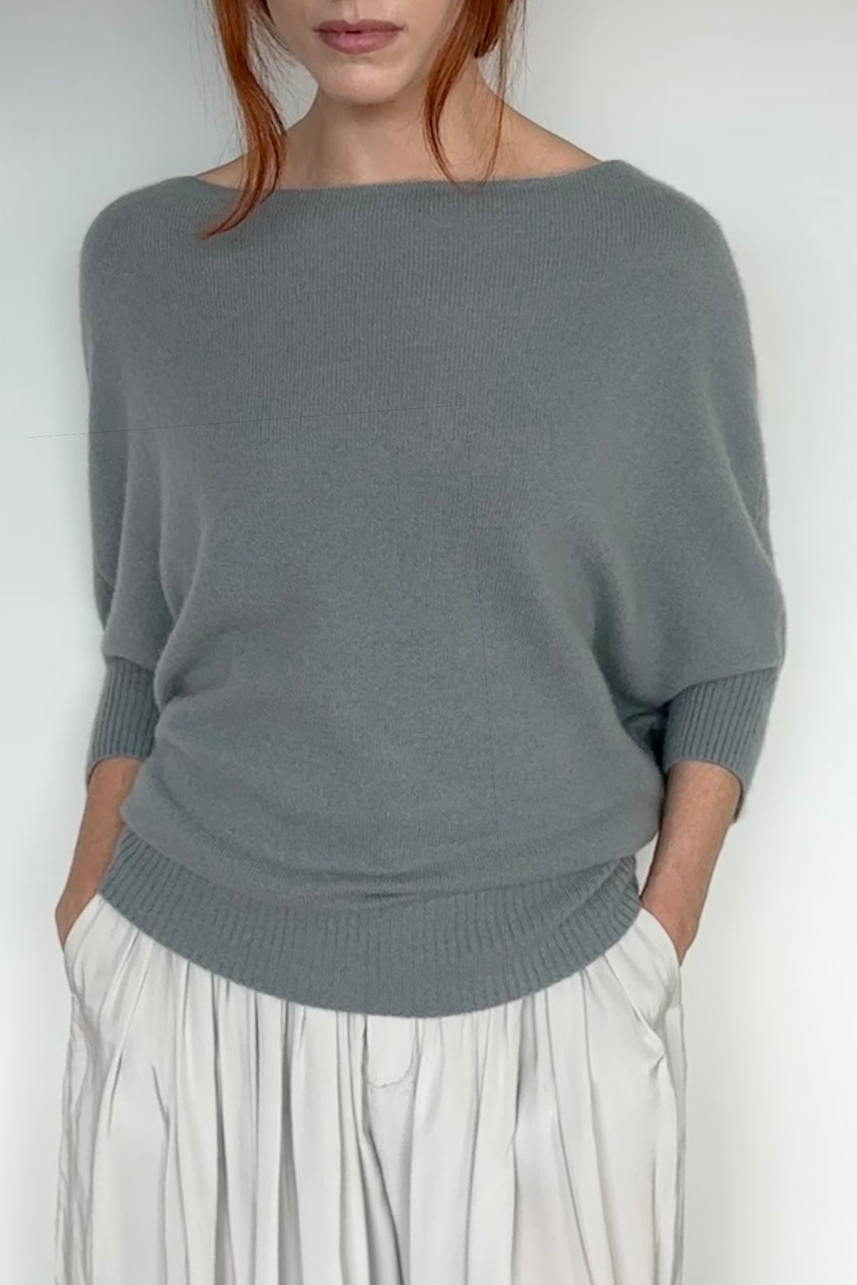 A woman wearing a slate gray knit sweater with a wide boat neckline and elbow-length sleeves, paired with light gray pleated trousers, against a plain white background.