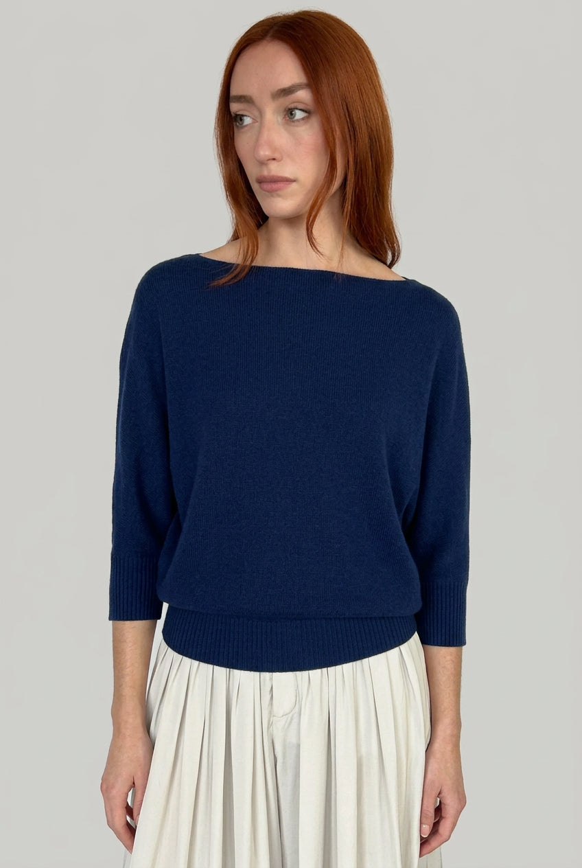 A woman with red hair wearing a cashmere navy knit sweater with a wide boat neckline and three-quarter sleeves, paired with a light gray pleated skirt, against a plain light gray background.