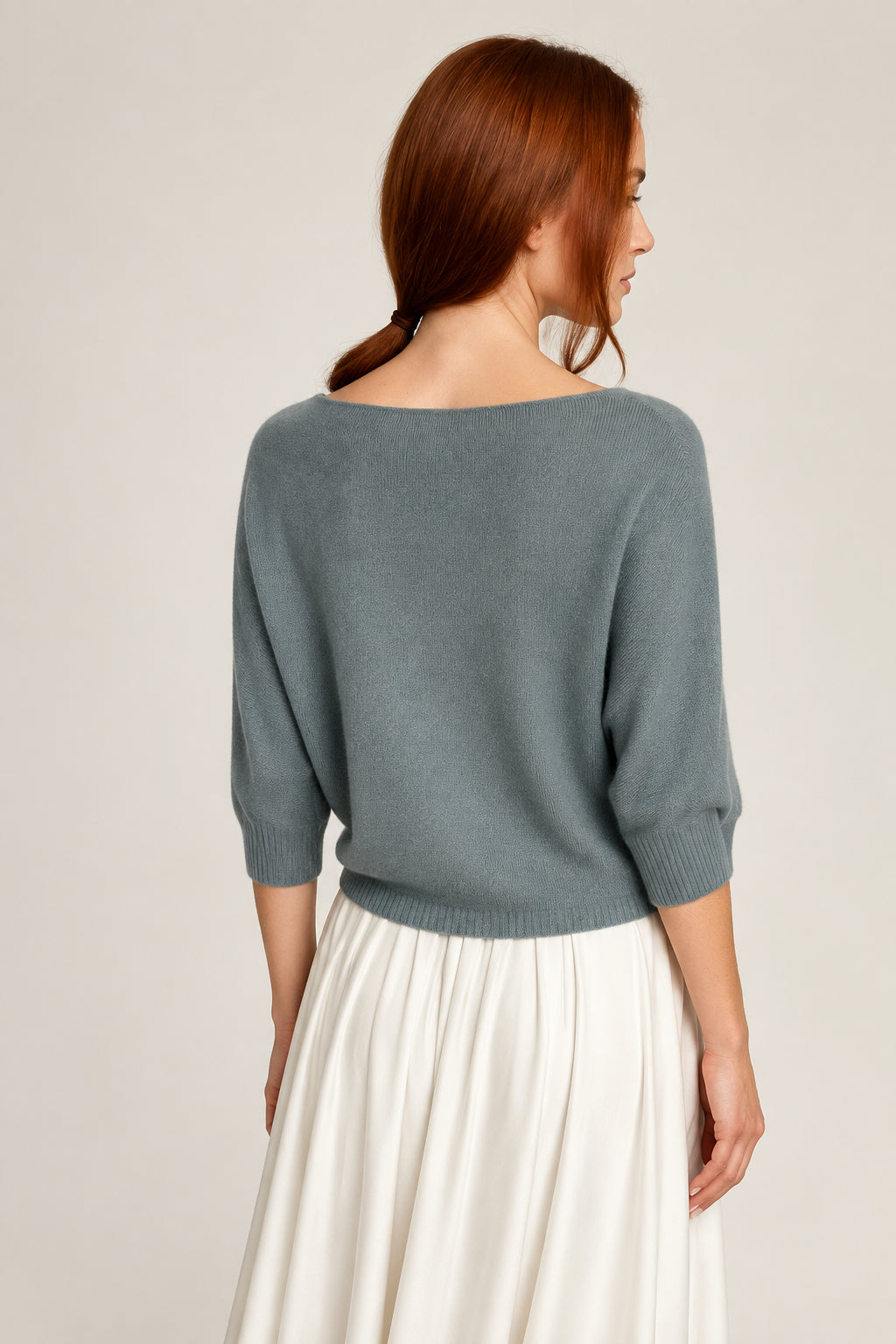 A woman with red hair is shown from the back wearing a slate blue-gray knit sweater with three-quarter sleeves and a wide boat neckline, paired with a white pleated skirt, standing against a plain light gray wall.