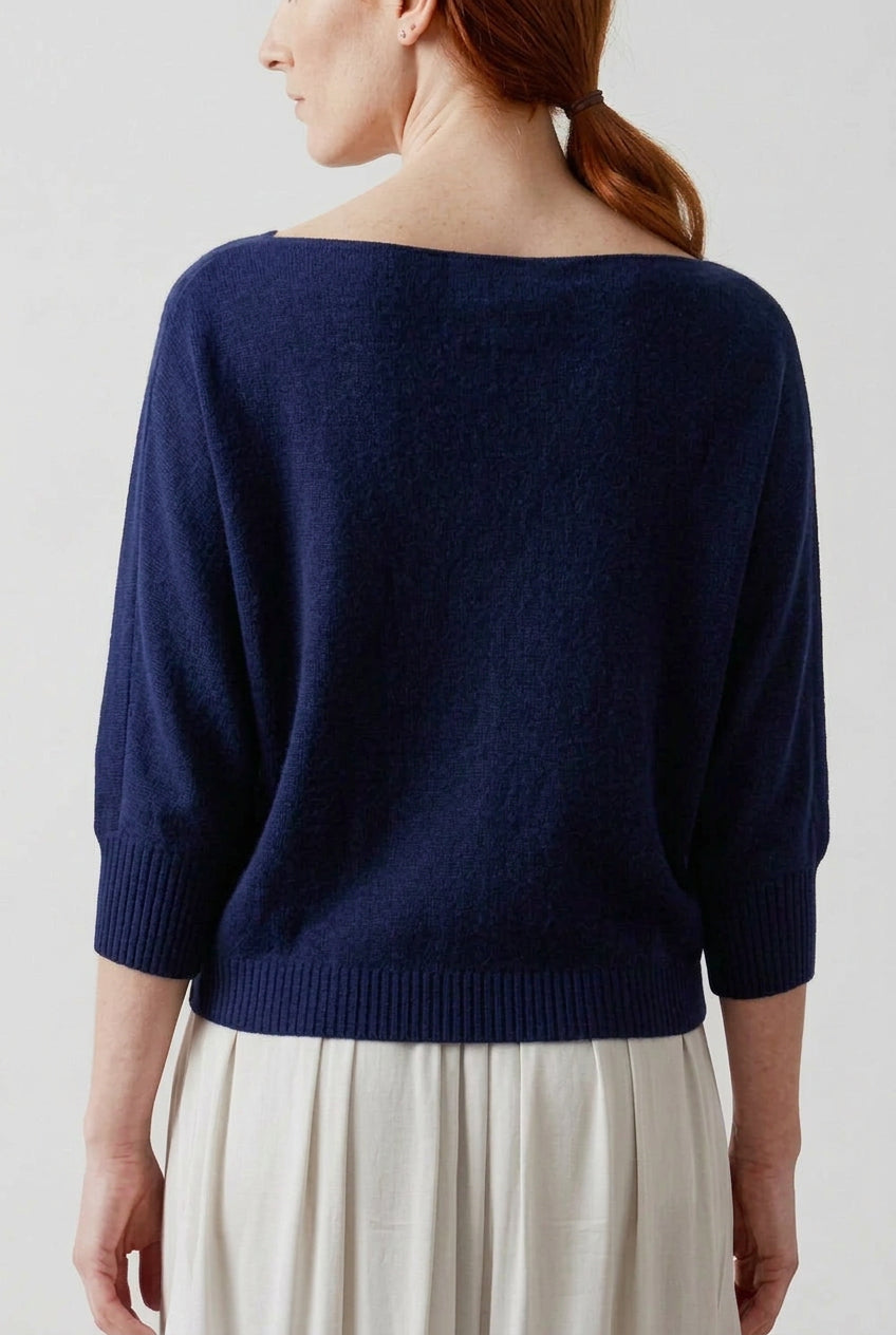 A woman with red hair tied back, shown from the back wearing a navy blue wide-neck knit sweater and cream-colored pleated skirt against a light gray studio background.