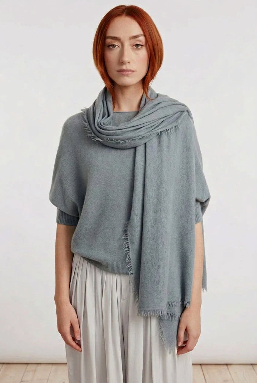 A woman with red hair stands against a plain light wall wearing a draped slate gray knit poncho over a light gray top and white pleated trousers.