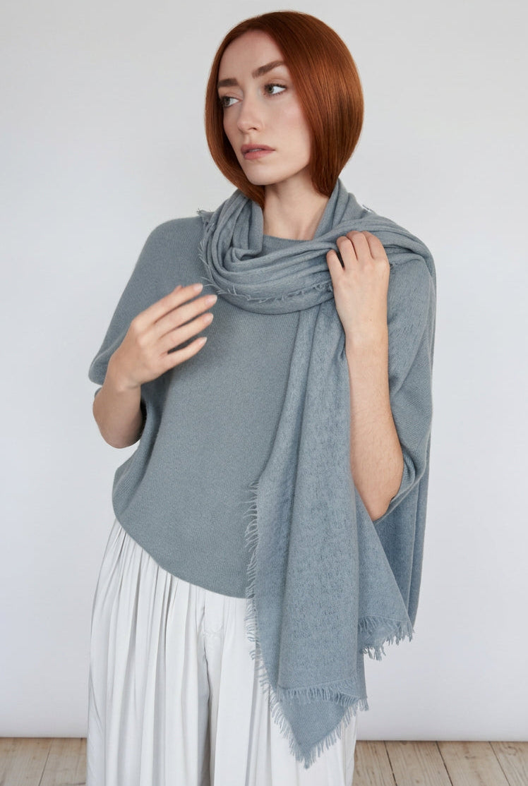 A woman with red hair models a light gray knit shawl draped over her shoulders, paired with a gray top and white pleated trousers, standing against a plain off-white wall.
