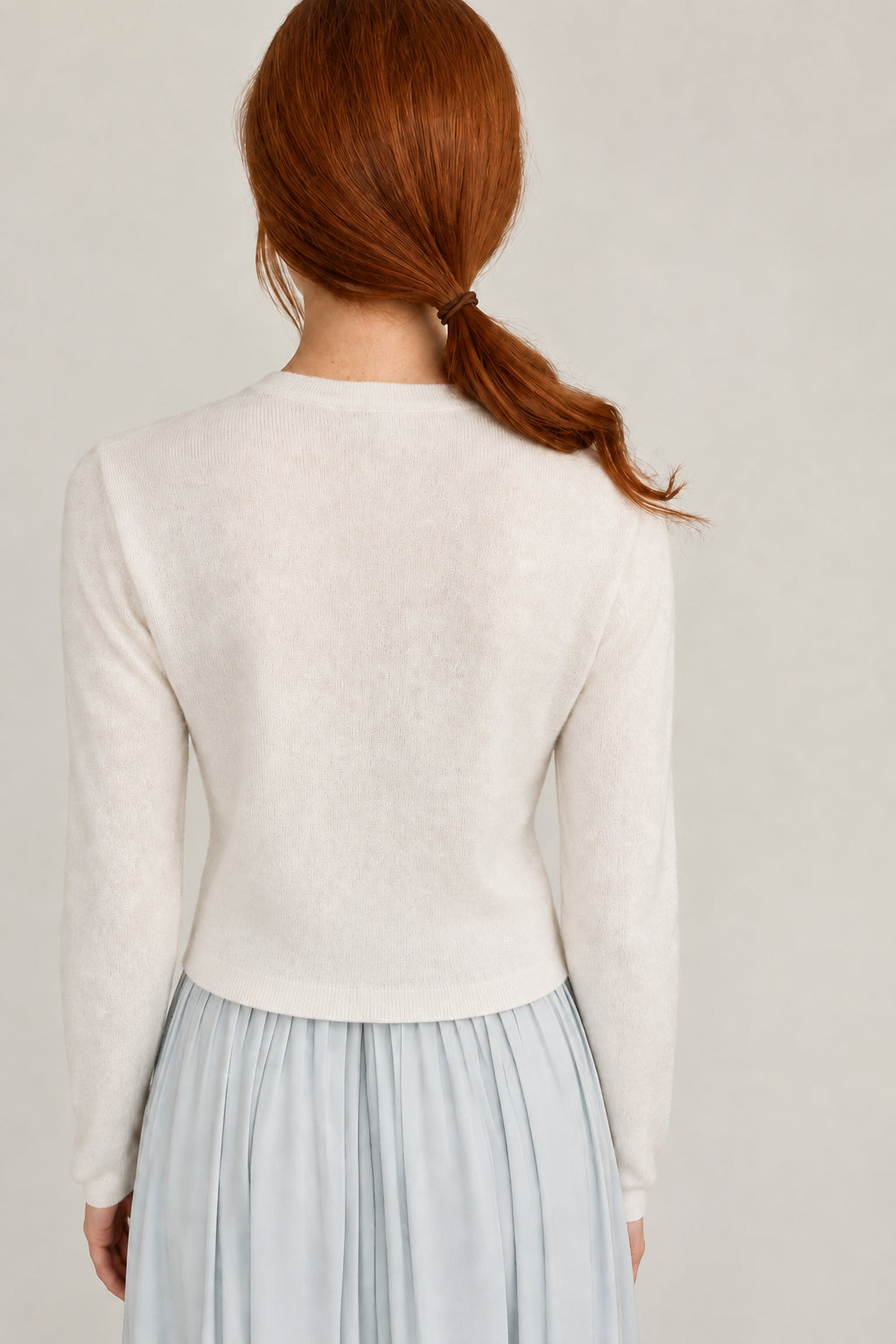 A person shown from behind wearing a light cream knit sweater and a pale blue pleated skirt, standing against a plain off-white wall.