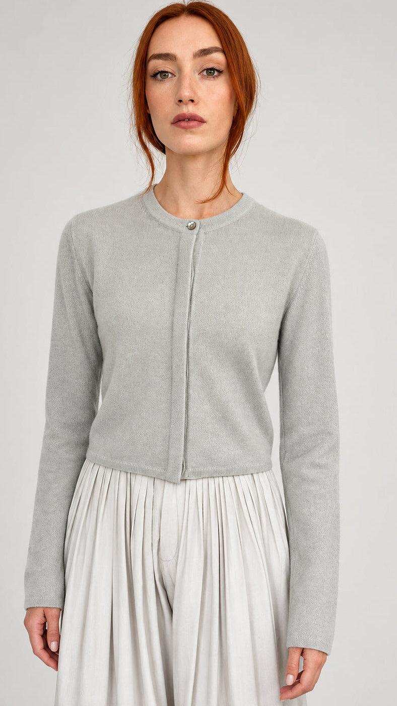 A woman with red hair wearing a light gray cashmere short cardigan and a matching light gray pleated skirt, standing against a plain off-white wall.