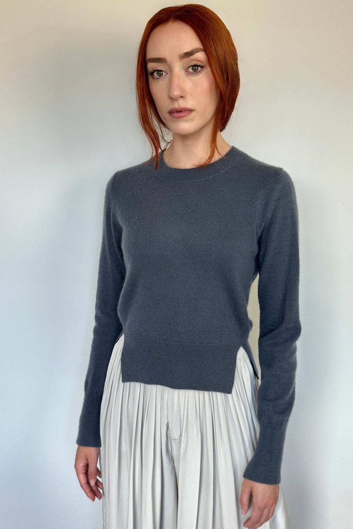 A woman with red hair wearing a dark slate gray long-sleeve knit sweater and a white pleated skirt, standing against a plain off-white wall.